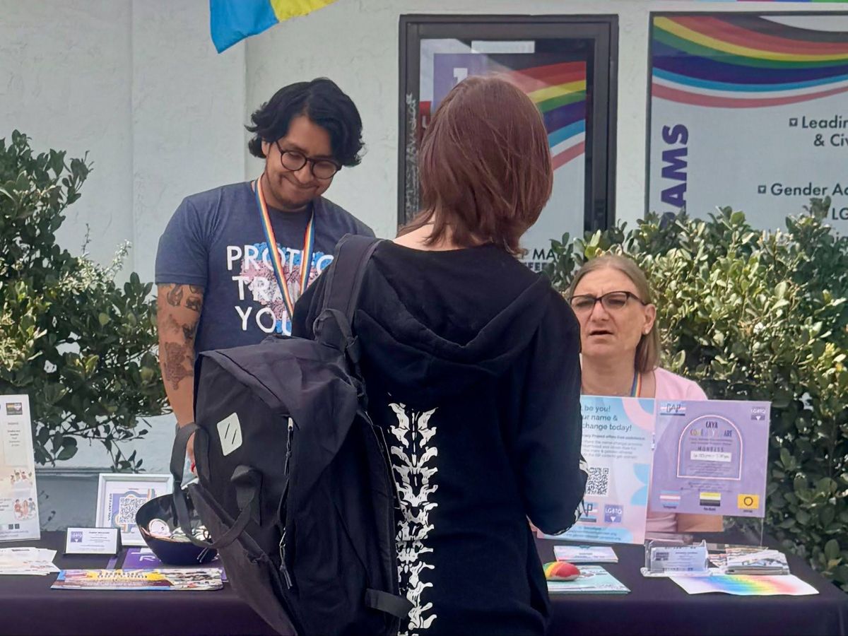 Two people, one standing and one sitting, talk to a young person with their back turned away from the camera in front of a table with multiple flyers and cards. 
