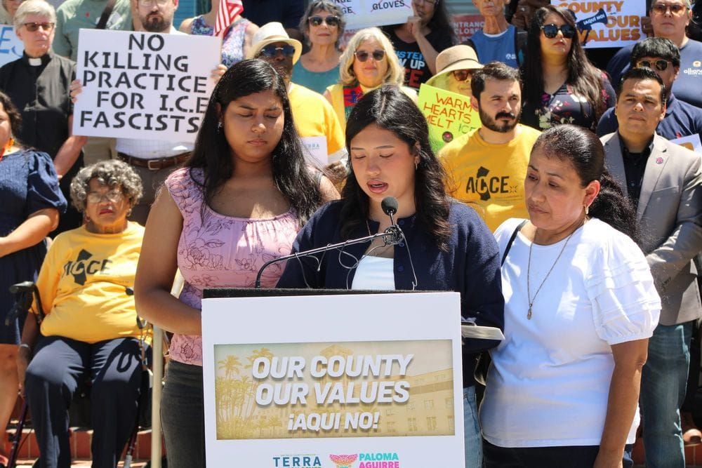 A group of three people stand at a podium with a group on stairs behind them, one holding a sign that read "No killing practice for ICE fascists."