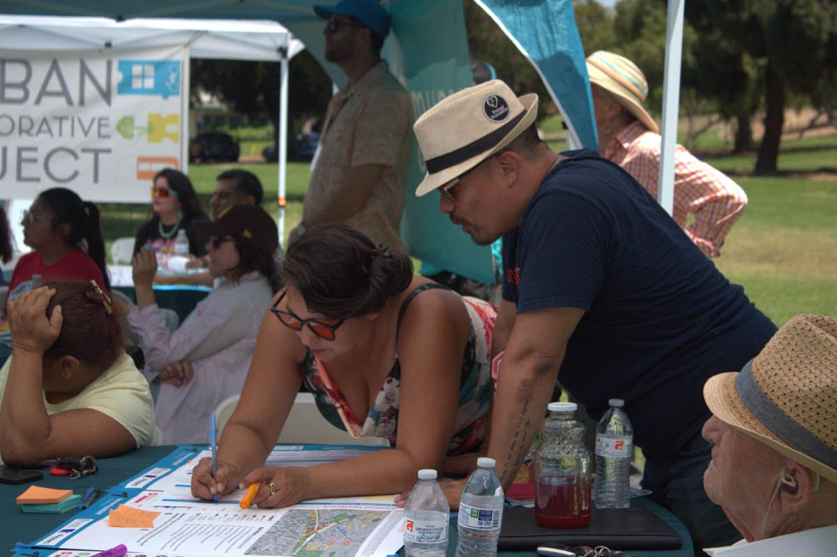 Multiple people in the background look to the left of the photo while two people hover over a map talking with one of them marking the map with a pen.