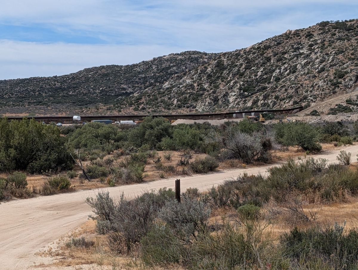 A rocky mountain with a rust brown wall running into it and construction equipment in front, surrounded by scrubby desert