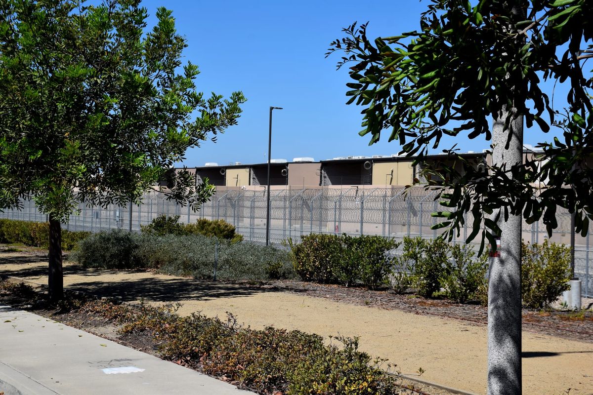 A prison building with a tall fence and razor wire with trees and a dirt path in front