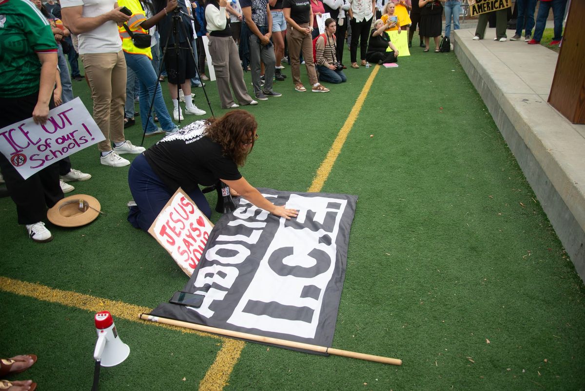 A person with long hair and a black shirt puts their hand on an Abolish I.C.E. flag that is lying flat on the ground