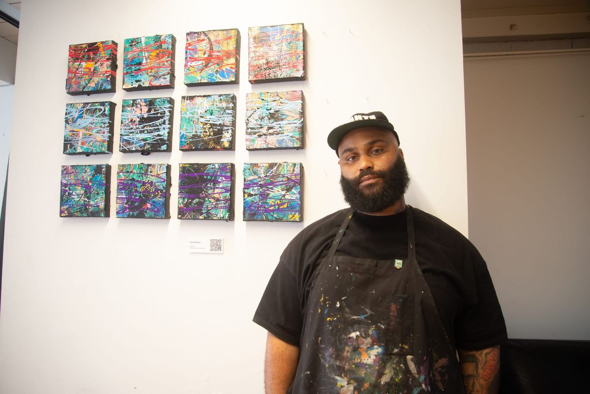 A man in a black cap, shirt and apron with paint on it stands in front of 12 square abstract paintings with a variety of colors arranged in rows of four