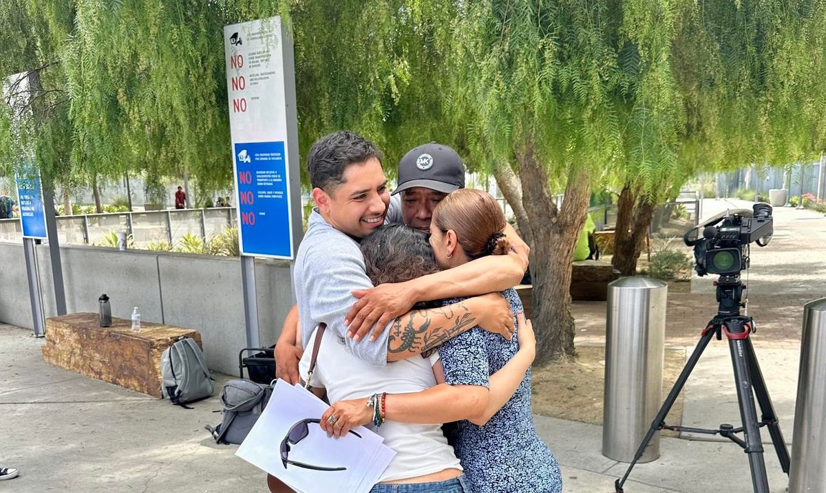Four people cling to each other in a close embrace while standing on a sidewalk with a tree and a tv camera in the background