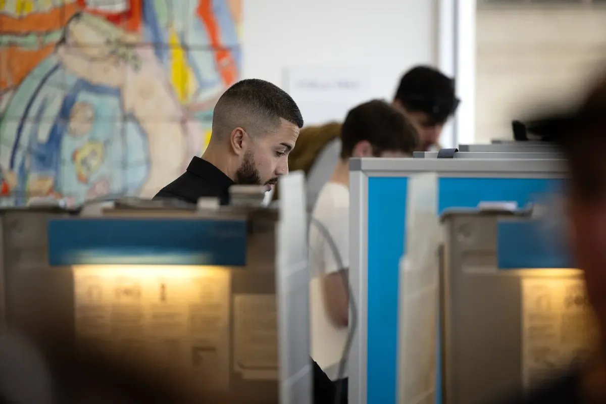 Picture of people filling out ballots in voting booths with a tiled mural on the wall behind them