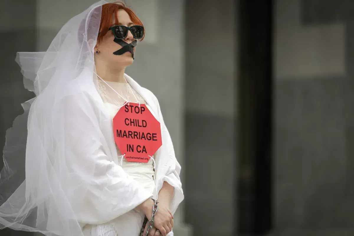 A person in a white wedding dress and veil stands outside a building with tape forming an "X" across their mouth, and a stop sign with the words "Stop Child Marriage in CA"