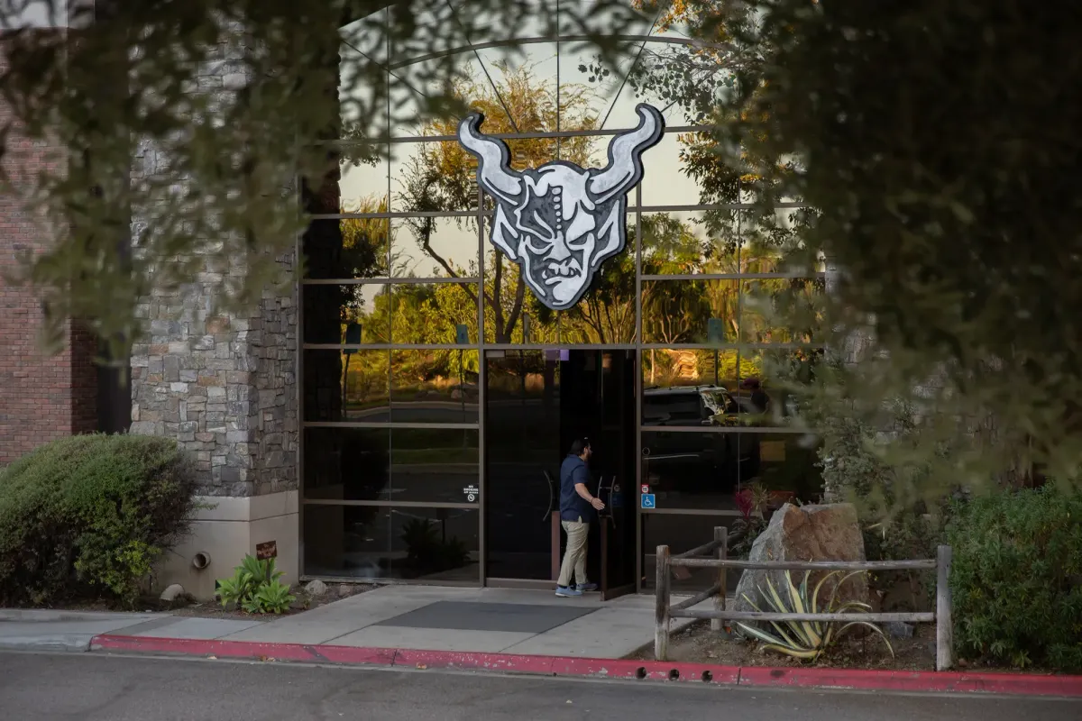 A person holds open a door to a building with a glass window front and the Stone Brewing symbol above the door