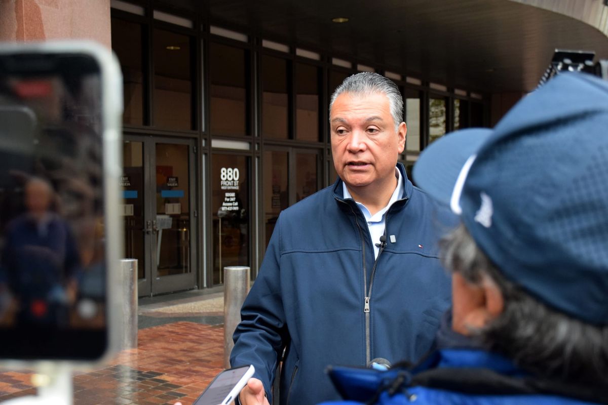 A man speaks outside in front of a building with some facing away from the camera to his right and a cellphone recording on the left
