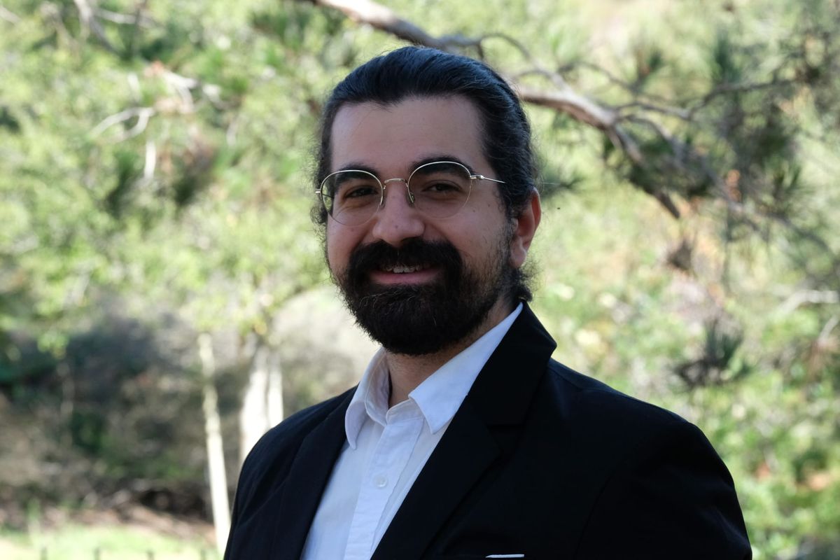 A person with a beard and glasses and wearing a suit stands in front of trees