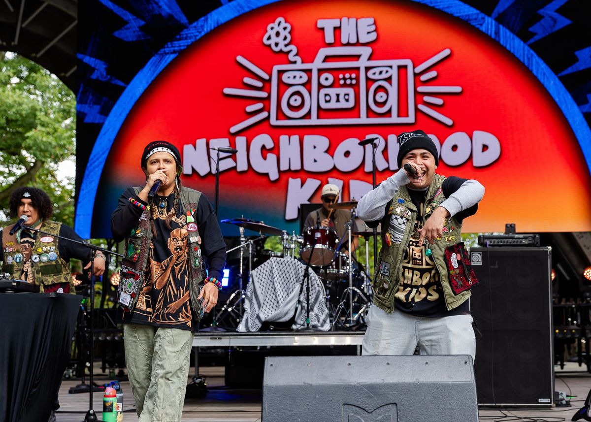 Two people wearing long sleeve shirts, vests and jeans hold microphones on a stage in front of a screen that says the neighborhood kids