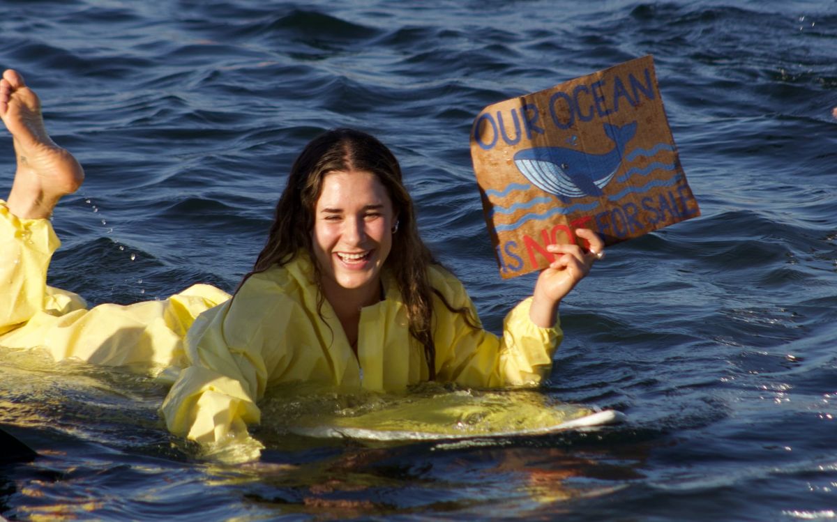 A person with long brown hair wearing a yellow hazmat suit lies on a surf board holding a sign that says our ocean is not for sale