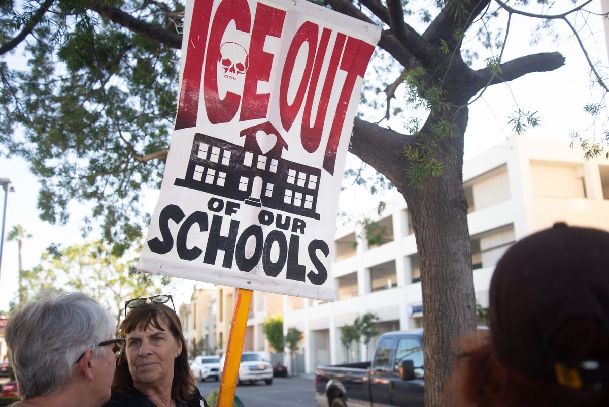 A person holds a red, white and black sign that says ICE Out of Our Schools standing in front of a tree and street