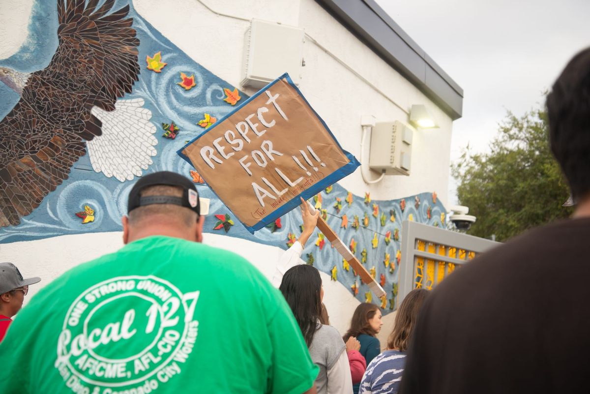 Several people, including one in a green shirt, walk away from the camera, on person holding a sign that says Respect For All. 