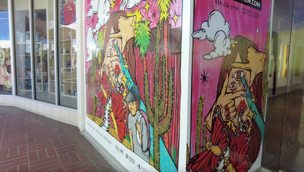 A printed poster spans to walls at an outdoor space with a drawing of a person wearing a backpack, butterflies a Cactus a person with a third eye with a beam of light coming out of it.