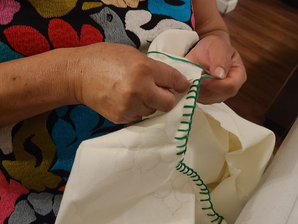 close up of a woman's hands sewing green thread on the edge of a white cloth