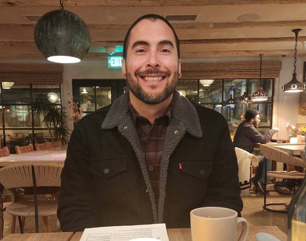 A man smiles sitting in a restaurant, wearing a black jacket