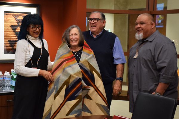 Four people, including one with a blanket wrapped around her, smile in a board room with water bottles on a table behind them