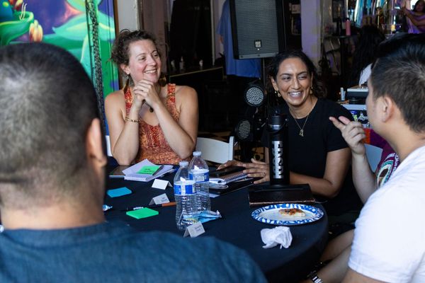 Two women smile at a table facing two people whose backs are to the camera. The table has water bottles and post-it notes on it.