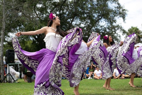 Four women wearing purple, patterned skirts, white shirts and flowers in their hair dance in front of a tree on the grass at an event with people sitting in the background