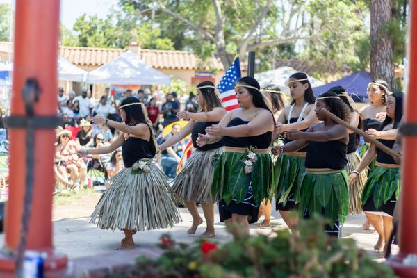 Dancers in grass and leaf skirts perform on a stage during an outdoor event with people in the background 