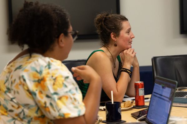A woman in a floral dress with her back to the camera looks at a woman in a green dress who looks off to the right. Both sit at a table with laptops and food in front of them.