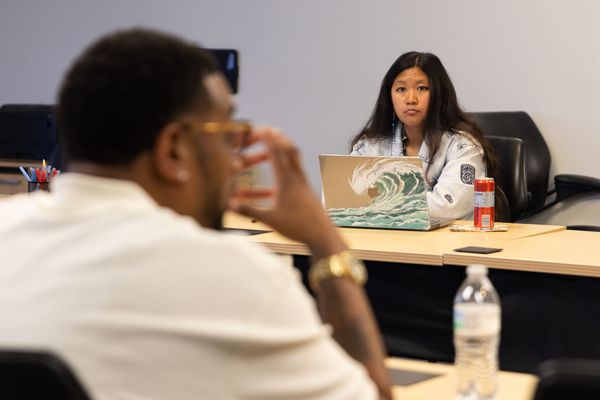 A woman faces the camera sitting at a table in front of a laptop with a man with his back faced to the camera in front of her