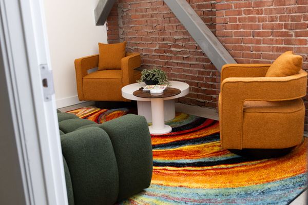 Two orange chairs and a green couch sit on a multi-colored rug in a small room with a white wall and a brick wall