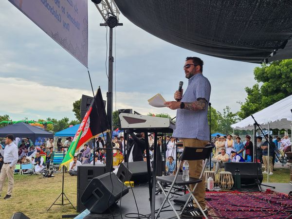 A man speaks on a stage at an outdoor festival with tents and people in the background