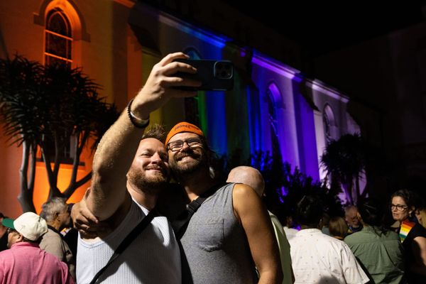 Two people with a crowd behind them take a selfie in front of a church with rainbow lights shining on it.