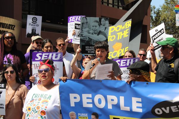 A person speaks into a microphone at a rally with other protesters nearby holding signs reading "Palantir," "Her husband was kidnapped in June", "Power to the People"