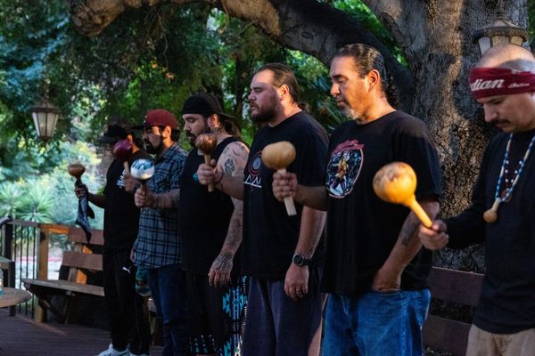six people stand under a tree singing songs and using gourd rattles