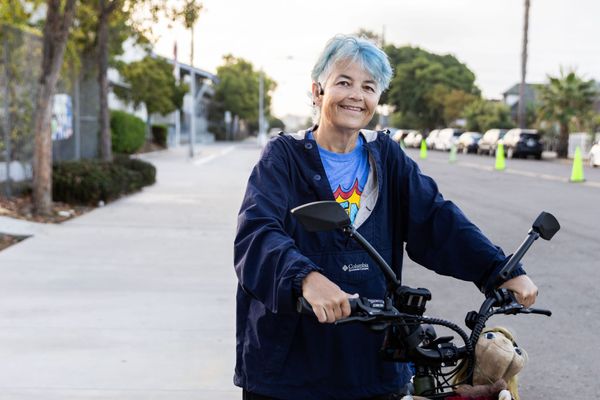 A person with blue hair and a blue jacket holds the handlebars of an e-bike with a school in the background