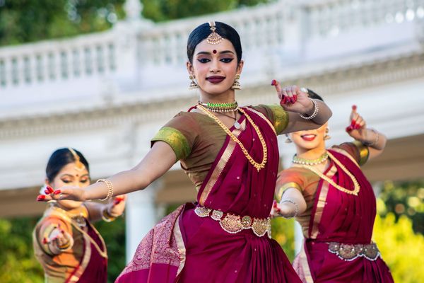 Three women dance while wearing red dresses and gold jewelry. Two women are in the background and the focus is on the woman in the center.