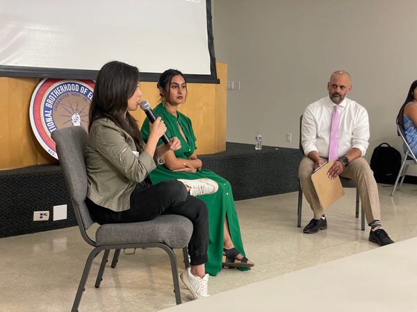 Three people sit in chairs on a panel at an event while one person speaks into a microphone