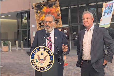 A man with a gray beard and dark suit stands next to another man in a suit in front of a podium that says U.S. House of Representatives