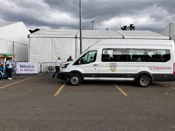 A white van with Gobernación written on the side is parked in front of a white tent with silhouettes of people leaving the van seen through the window