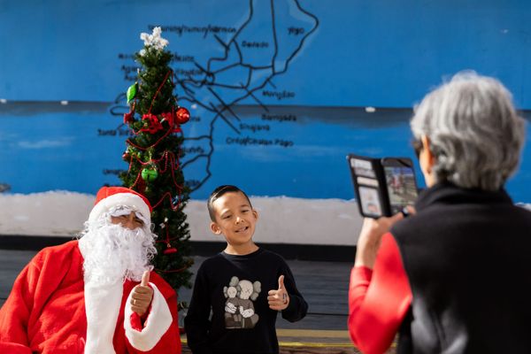 A man and a child stand next to each other holding a thumbs up for a woman holding her phone to take a photo of them.