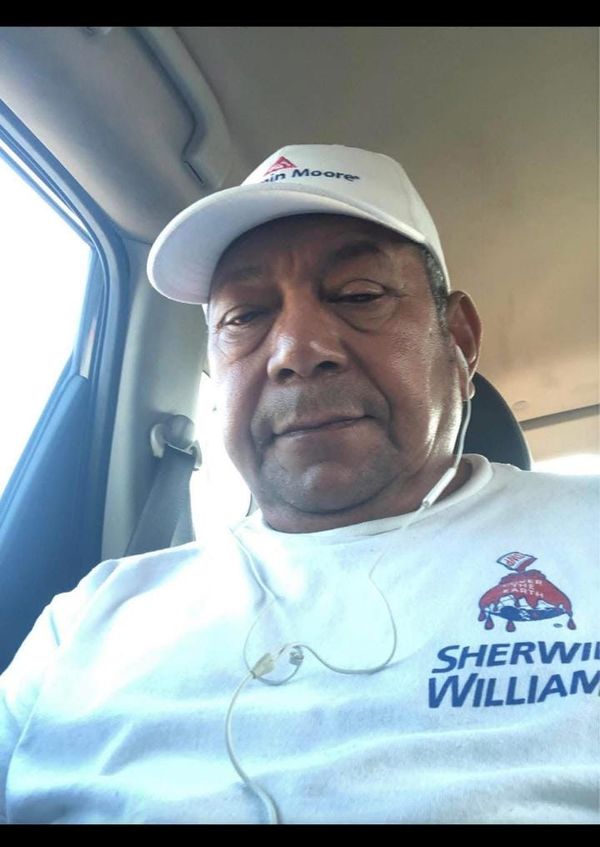 A man in a white shirt and hat sits in a car taking a selfie