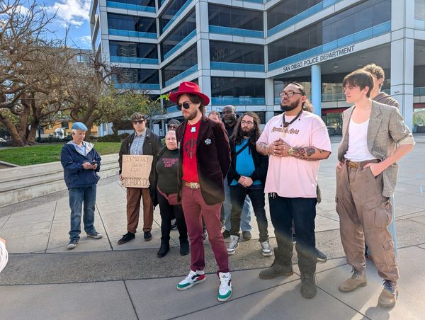 A man in red clothing and a hat stands at the front of a group of people in a plaza in front of a building with a sign that says San Diego Police Department