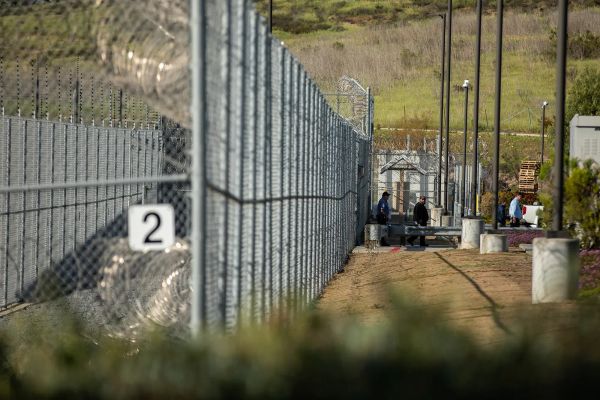 Two people walk out of a gate in a tall metal fence with razor wire on top