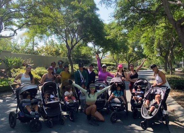 A group of women with children and strollers pose in front of a tree-covered trail.