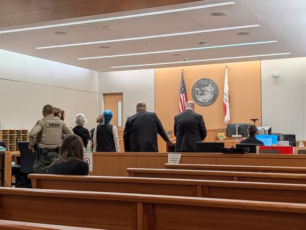 a court room with several women and two men standing facing away from the camera and a bailiff behind them with a sheriff deputy uniform on