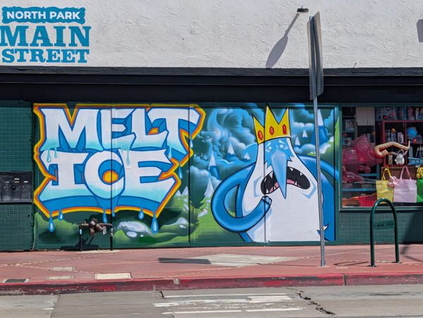 A green wall with a painted message of "Melt ICE" in white, blue, orange and yellow with a blue character with a crown and white beard standing next to it
