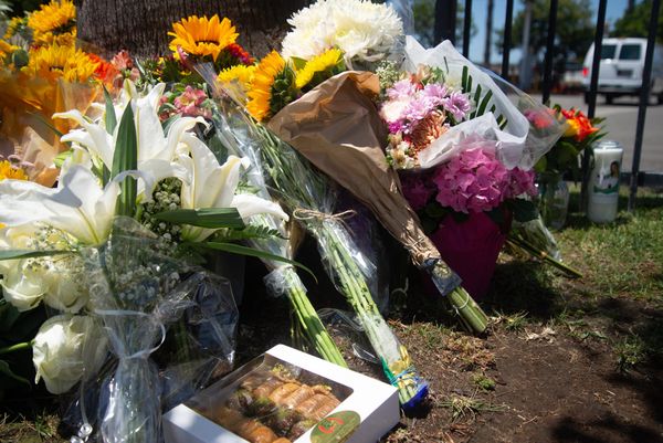 Bouquets of lilies and sunflowers lean against a palm tree and a box of baklava lies on the ground
