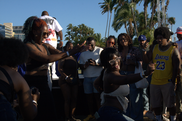 People stand in a circle and dance outside in front of palm trees