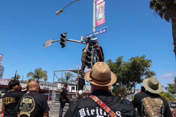 Close up of the back of people standing watching someone on scaffolding hang up a sign on a street light with a blue sky in the background