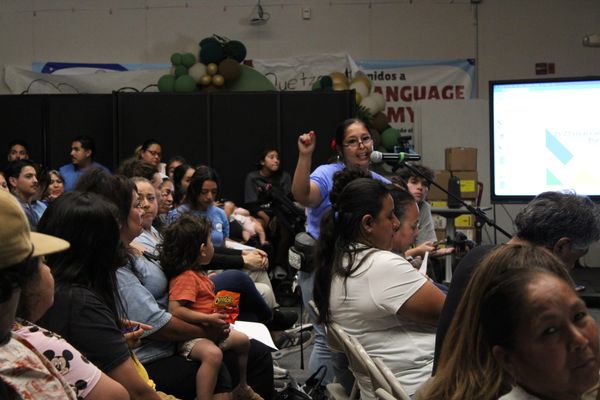 A woman speaks into a microphone while holding her hand up and other people in the audience watch