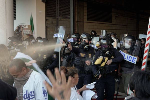Department of Homeland Security officials in riot gear cluster together while one sprays an orange-ish substance at protesters who are standing close to them