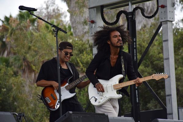 One person plays a bass guitar and another plays the guitar on stage in front of trees at an outside music festival