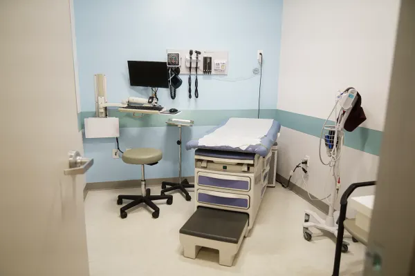 A medical exam table, computer, stool and blood pressure machine are in a small room with a white wall and a blue wall